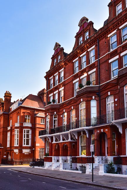 A street scene showing a large, multi-storey red brick building with white window frames and decorative architectural details in West Kensington. The ground floor features a covered entrance with white wooden supports and steps leading up to a door. Adjacent buildings of similar style can be seen along the street under a clear blue sky. In the foreground, a black removal van from Removal Van West Kensington is parked on the pavement, with its rear doors open, ready for a home relocation process. Inside the van, various moving materials including cardboard boxes, wrapped furniture pieces in plastic and fabric, and protective blankets are visible, with some items being carried or loaded onto the vehicle using trolleys and straps. The scene captures the preparations involved in furniture transport and packing during a professional removal or moving service, illustrating the logistical aspects of house removals and the organization of belongings for efficient transport.