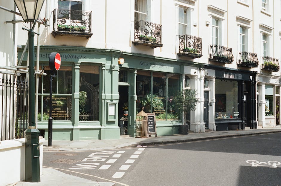 A street scene showing the ground floor of a white multi-story building with several black wrought iron balconies, each decorated with potted plants and flowers. The building's ground level features a flower shop with a light green facade, displaying large shop windows filled with plants and floral arrangements. An A-frame chalkboard sign and a birdcage-style flower stand are positioned outside the shop. A sidewalk runs in front of the shop, with a traffic sign and a black metal pole at the corner, and a designated bike lane marked on the road. The image depicts the area during daytime with natural lighting, illustrating an urban setting suitable for house removals or home relocation services, with [COMPANY_NAME] potentially providing furniture transport and packing assistance in West Kensington.