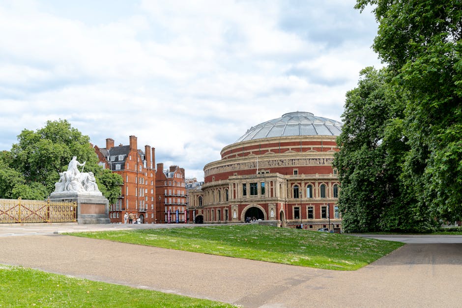 A wide view of a city park with a large, historic circular building featuring a domed roof and arched windows, situated behind a grassy area and paved pathways. To the left, there is a white marble sc