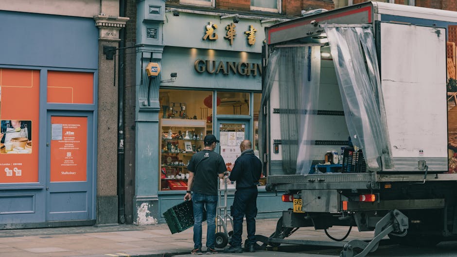 Two movers from Removal Van West Kensington are preparing for a home relocation outside a shop called 'Guang' near Kensington Olympia station. One mover, wearing a black cap and dark clothing, is holding a plastic crate, while the other, dressed in a dark jacket, stands nearby with their back to the camera. They are next to a large moving truck parked on the pavement, with its rear ramp open and covered with clear plastic sheeting, revealing the interior with packing materials and boxes. In front of the shop, there are visible window displays and a blue door with orange accents, along with signage above the entrance. The scene is set during daylight with natural lighting, and the environment suggests a busy urban setting involved in furniture transport, packing, and the logistics of a commercial removal, capturing the preparation process for a shop's move near Kensington Olympia station.
