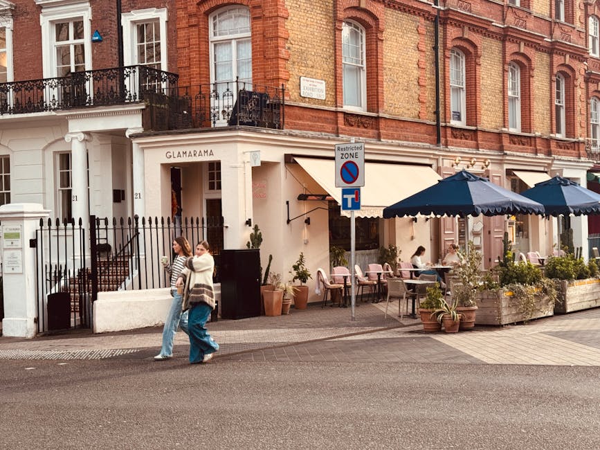 Two movers from Removal Van West Kensington are preparing for a home relocation outside a shop called 'Guang' near Kensington Olympia station. One mover, wearing a black cap and dark clothing, is holding a plastic crate, while the other, dressed in a dark jacket, stands nearby with their back to the camera. They are next to a large moving truck parked on the pavement, with its rear ramp open and covered with clear plastic sheeting, revealing the interior with packing materials and boxes. In front of the shop, there are visible window displays and a blue door with orange accents, along with signage above the entrance. The scene is set during daylight with natural lighting, and the environment suggests a busy urban setting involved in furniture transport, packing, and the logistics of a commercial removal, capturing the preparation process for a shop's move near Kensington Olympia station.