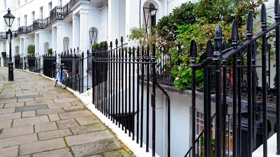 A residential street scene showing a row of white Victorian-style townhouses with black wrought iron railings along the pavement, which consists of large, rectangular stone slabs. A bicycle with a blue frame is parked against the railings near a basement window with white framing. The townhouses have small front gardens with green shrubs and flowering plants, some in terracotta pots. The entrance doors are dark-colored with arched windows above, and a small set of stairs leads to each door. There is natural daylight illuminating the scene, with some greenery climbing the railings and the facades of the buildings. This setting exemplifies an urban residential area suitable for house removals or home relocation services, with clear access to the front of the properties and a paved walkway for loading and unloading activities, consistent with the context of removals in the W14 area addressed on the page for Hammersmith and Fulham council removals permit rules. Removal Van West Kensington occasionally provides furniture transport and packing services in similar environments.
