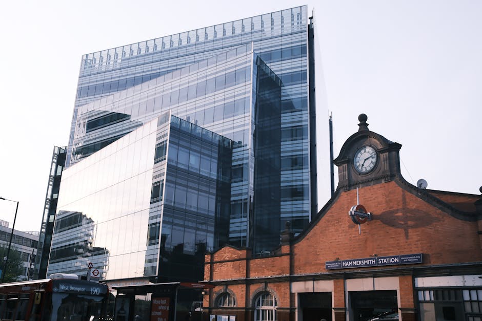 A modern glass office building with reflective windows towering above a traditional brick railway station building, which has a clock and a sign reading 'Hammersmith Station'. The photo captures the loading area where a removal van from Removal Van West Kensington is stationed nearby. Several cardboard boxes, some wrapped in plastic wrap, are seen on the pavement and around the entrance, likely prepared for a home relocation or furniture transport. A trolley is present, suggesting ongoing loading or unloading activities, with an area that appears designated for moving services, such as packing, lifting, or carrying items from the station to the vehicle. The lighting indicates daytime with an overcast sky, providing natural light that illuminates all objects clearly. The scene reflects typical logistics involved in house removals and transportation, aligned with regulations for removals permits around Hammersmith and Fulham W14 area.