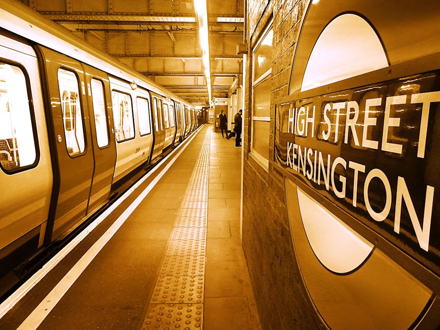Inside the London Underground station at High Street Kensington, the platform is lined with beige tiles and a tactile paving strip along the edge. A yellow Northern line tube train is stationary on the left side, with multiple closed windows and doors. On the right wall, a large circular sign features the station name 'High Street Kensington' in white text on a black background, displayed within the iconic roundel symbol. The station's brick wall behind the sign is visible, with some lighting fixtures illuminating the platform. In the distance, a few passengers are waiting, standing near the platform edge. The scene depicts a typical home relocation environment where furniture and boxes are transported onto or off the train during a packing and moving process, facilitated by [COMPANY_NAME], specialists in removals and loadings in West Kensington.