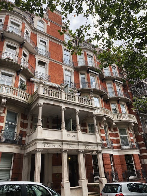 A multi-storey residential building with a red brick facade and decorative white stone architectural features, including curved balconies with metal railings and ornate corbels. The building's entrance is supported by white stone columns and has a sign reading 'CAMPDEN HILL COURT' above the doorway. In the foreground, part of a city pavement is visible with parked cars along the street. The scene is framed by overhanging green-leaved trees, with natural daylight illuminating the building's exterior, providing context for house removals or furniture transport activities associated with relocation services.
