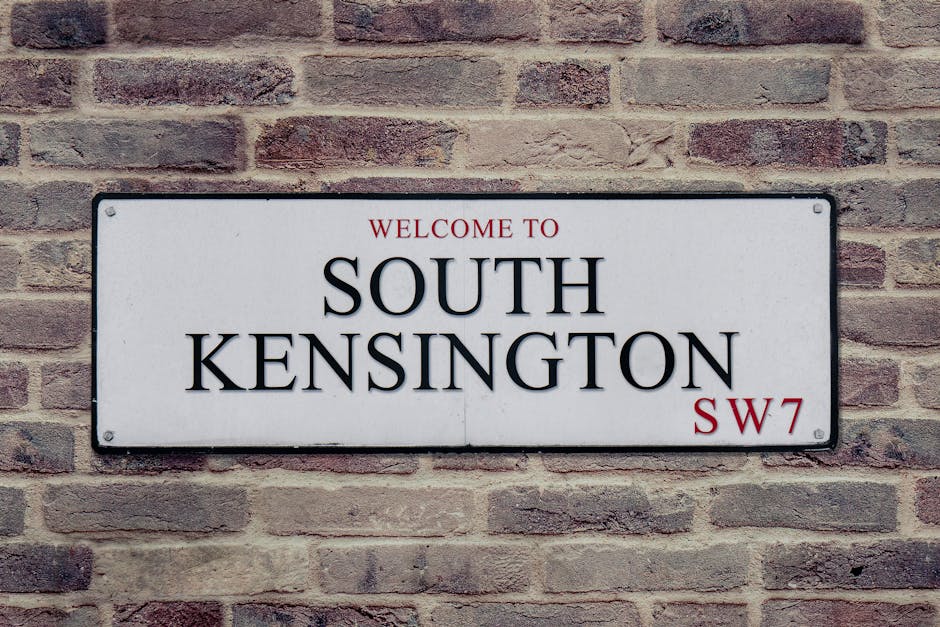 Inside the London Underground station at High Street Kensington, the platform is lined with beige tiles and a tactile paving strip along the edge. A yellow Northern line tube train is stationary on the left side, with multiple closed windows and doors. On the right wall, a large circular sign features the station name 'High Street Kensington' in white text on a black background, displayed within the iconic roundel symbol. The station's brick wall behind the sign is visible, with some lighting fixtures illuminating the platform. In the distance, a few passengers are waiting, standing near the platform edge. The scene depicts a typical home relocation environment where furniture and boxes are transported onto or off the train during a packing and moving process, facilitated by [COMPANY_NAME], specialists in removals and loadings in West Kensington.
