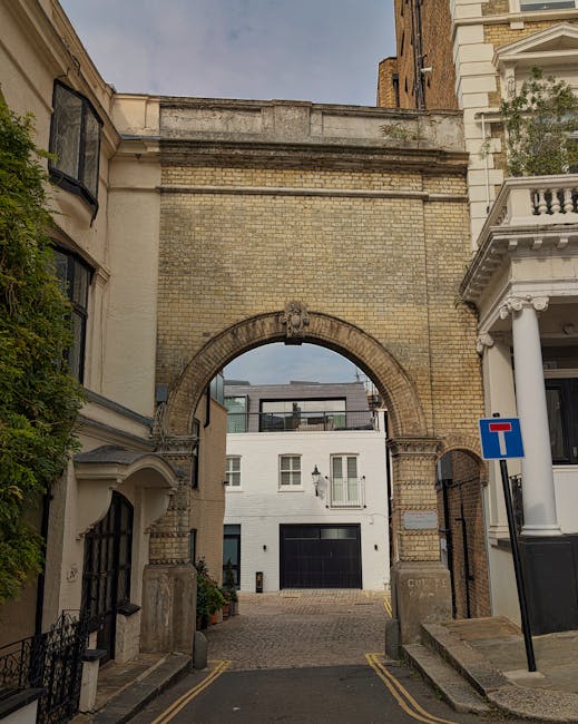 The image depicts a narrow urban street leading through a historic brick archway that connects two buildings, with a modern white residential building visible beyond the arch. The street has cobblestone paving and is framed by traditional terraced and Victorian-style architecture, including a building with white columns and decorative mouldings on the right. A blue and red 'no entry' traffic sign is mounted on a pole to the right side of the street. The scene is captured in daylight with natural lighting, and greenery is visible on the left side, suggesting a residential area in West Kensington. The image may relate to house removals or furniture transport services conducted by Removal Van West Kensington, especially in properties with narrow access points for efficient home relocation and moving logistics.