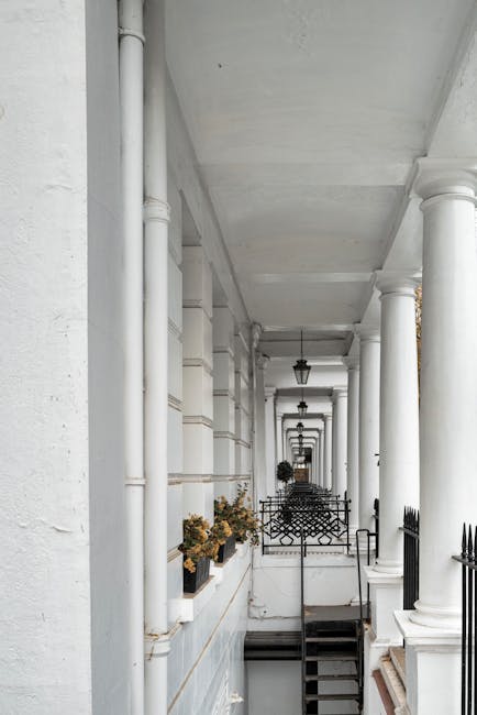 A long, narrow exterior corridor of a residential building in West Kensington features white painted walls and large, classical-style white columns supporting a ceiling with hanging black lantern-style light fixtures. On the left side, several window planters contain dried flowers, while on the right side, a series of small black metal fence panels and black handrails guide towards an open doorway at the far end. The pavement is partly visible, and a small set of steps leads down to a lower level. The scene depicts a house remodelling or home relocation process, with the corridor prepared for furniture transport or moving activities by Removal Van West Kensington, as part of their house removals service for properties with limited access.