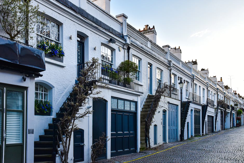 The image depicts a narrow urban street leading through a historic brick archway that connects two buildings, with a modern white residential building visible beyond the arch. The street has cobblestone paving and is framed by traditional terraced and Victorian-style architecture, including a building with white columns and decorative mouldings on the right. A blue and red 'no entry' traffic sign is mounted on a pole to the right side of the street. The scene is captured in daylight with natural lighting, and greenery is visible on the left side, suggesting a residential area in West Kensington. The image may relate to house removals or furniture transport services conducted by Removal Van West Kensington, especially in properties with narrow access points for efficient home relocation and moving logistics.