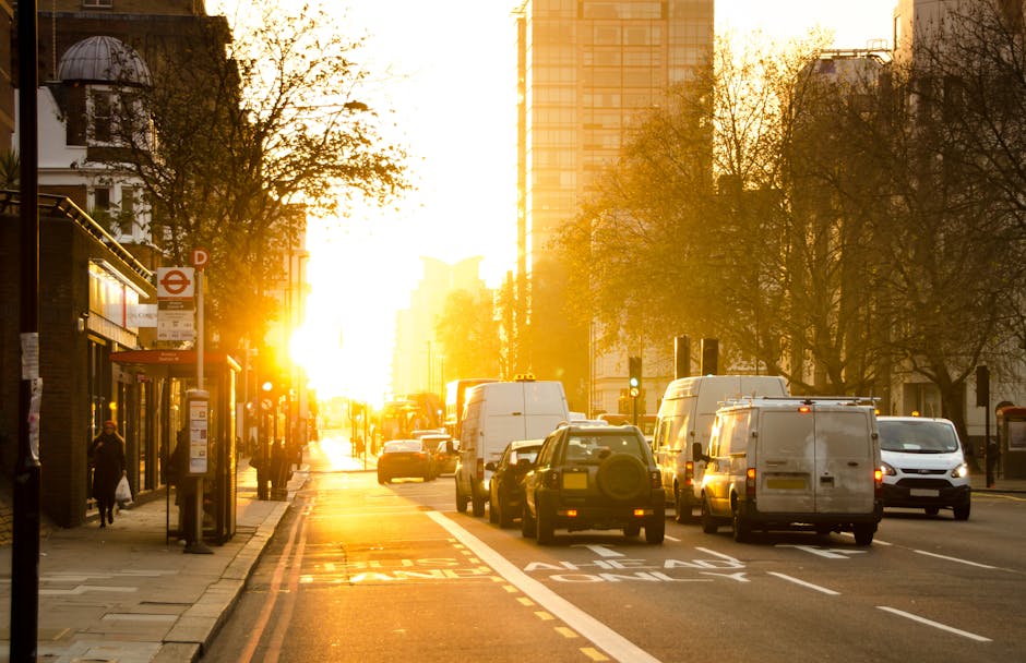 A busy city street during sunset with vehicles parked and moving along the roadside; in the foreground, a team from Removal Van West Kensington is seen loading furniture and packed boxes into a white moving van, which is partially visible on the right side of the image. The furniture includes cardboard boxes, some wrapped in plastic, and wooden furniture pieces. The scene is set next to a pavement with a bus stop and a few pedestrians nearby, all illuminated by the warm glow of the setting sun, casting long shadows across the street. Tall buildings and leafless trees line the background, indicating an urban environment typical for house removals and home relocation services in West Kensington. The overall atmosphere captures the process of packing and moving involving furniture transport and loading activities, reflective of a professional same-day removals operation to avoid last-minute delays, as provided by Removal Van West Kensington.