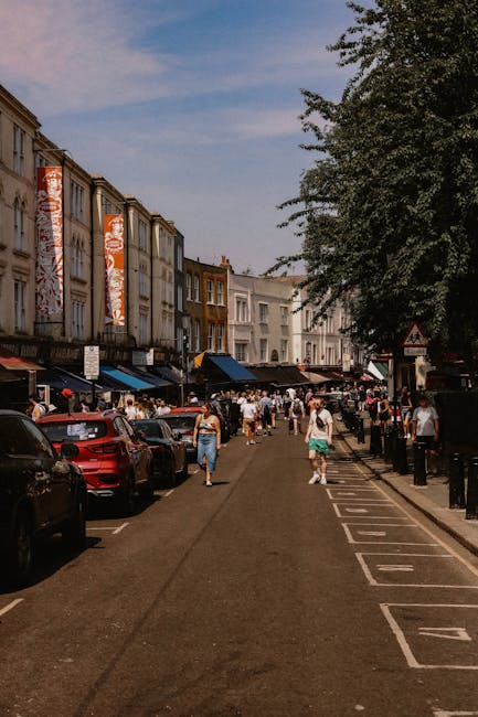 The image depicts a busy street scene in a residential area, with multi-storey terraced houses lining the left side of the road. The houses have white and beige facades, some with visible windows and doorways. A row of parked cars, predominantly black, gray, and red, lines the left curb. Pedestrians are walking along the street, with a few individuals engaged in activities related to home relocation, such as carrying cardboard boxes and small packages. On the right side of the street, there is a designated parking zone marked with white lines and black bollards separating the parking area from the pedestrian walkway. A large green tree extends over the sidewalk, providing shade. The sky is partly cloudy with patches of blue visible. This scene is suitable for illustrating elements of furniture transport, packing, and moving logistics, with a natural focus on the urban environment typical of house removals services provided by companies like Removal Van West Kensington.
