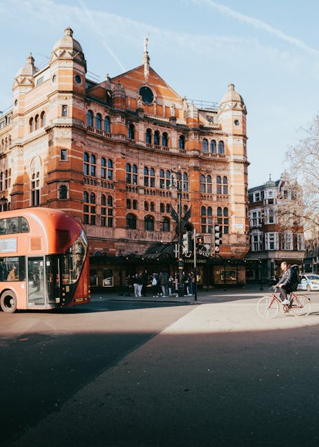 The image depicts a busy urban street scene outside a large, historic red-brick building with ornate architectural details, including rounded turrets and decorative window arches, situated in West Kensington. In the foreground, a red double-decker bus is stopped, with passengers visible inside and on the pavement nearby. A cyclist wearing a helmet and dark clothing rides along the road to the right. Pedestrians, some carrying shopping bags, are walking on the sidewalk adjacent to the building, which features a ground-floor commercial space with glass windows. The sky above is clear with some wispy clouds. This scene illustrates a typical day in West Kensington, capturing lively street activity, transportation, and the historic architecture, consistent with the type of residential and commercial environment served by Removal Van West Kensington's house removal and relocation services like packing, furniture transport, and loading processes.