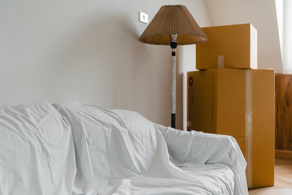 Indoor scene showing a living room corner with a white sofa covered in a protective sheet, positioned along a light grey wall. Behind the sofa, there are three cardboard moving boxes stacked next to a wooden cabinet, some with visible tape. To the left of the boxes, a tall floor lamp with a brown pleated lampshade stands on a black pole, illuminating the area. The environment suggests a home interior prepared for a relocation process, with furniture and packing materials in place, likely during the packing or moving stage in a house removal. The space is well-lit with natural light entering from an unseen window, and the setting reflects typical packing and furniture transport preparations associated with removals by companies such as Removal Van West Kensington, especially when considering potential hidden charges and detailed moving logistics.